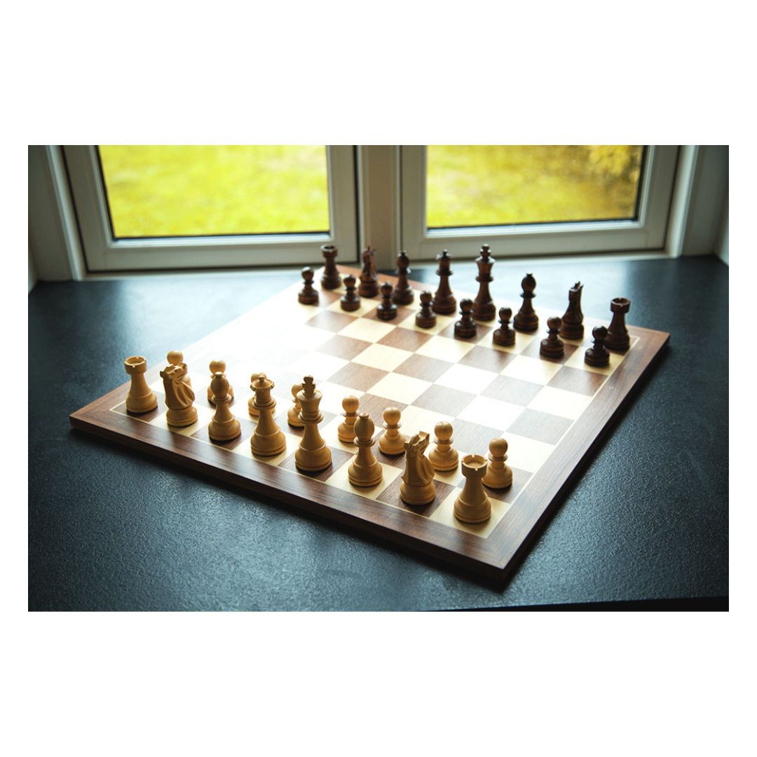 
Schackspel i trä som står på en mörk yta i en fönsterkarm med utsikt över en trädgård, där ljuset kommer in och gör att schackspelet kastar skuggor